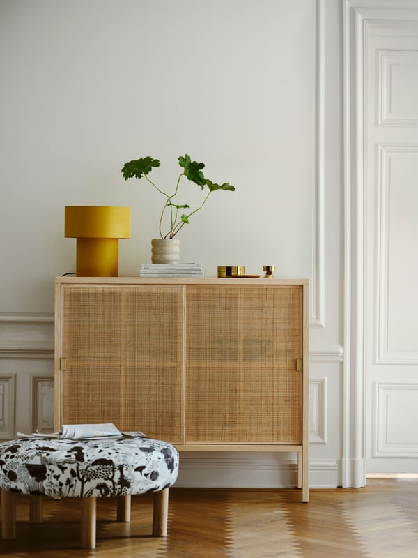 A STOCKHOLM 2025 cabinet with sliding doors in rattan and pine on parquet, with a table lamp on top and a pouffe in front.