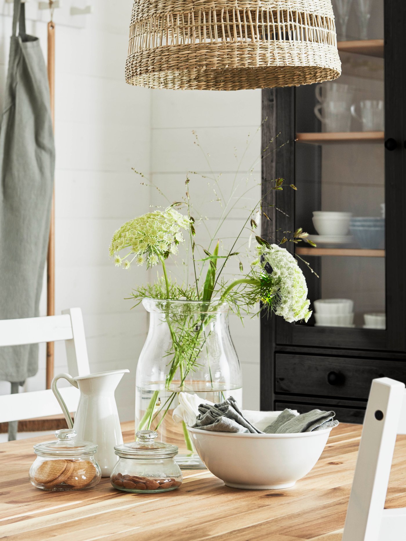 Un jarrón con flores y tallos, un cuenco para servir y un jarrón blanco sobre una mesa de comedor. Detrás hay una vitrina.
