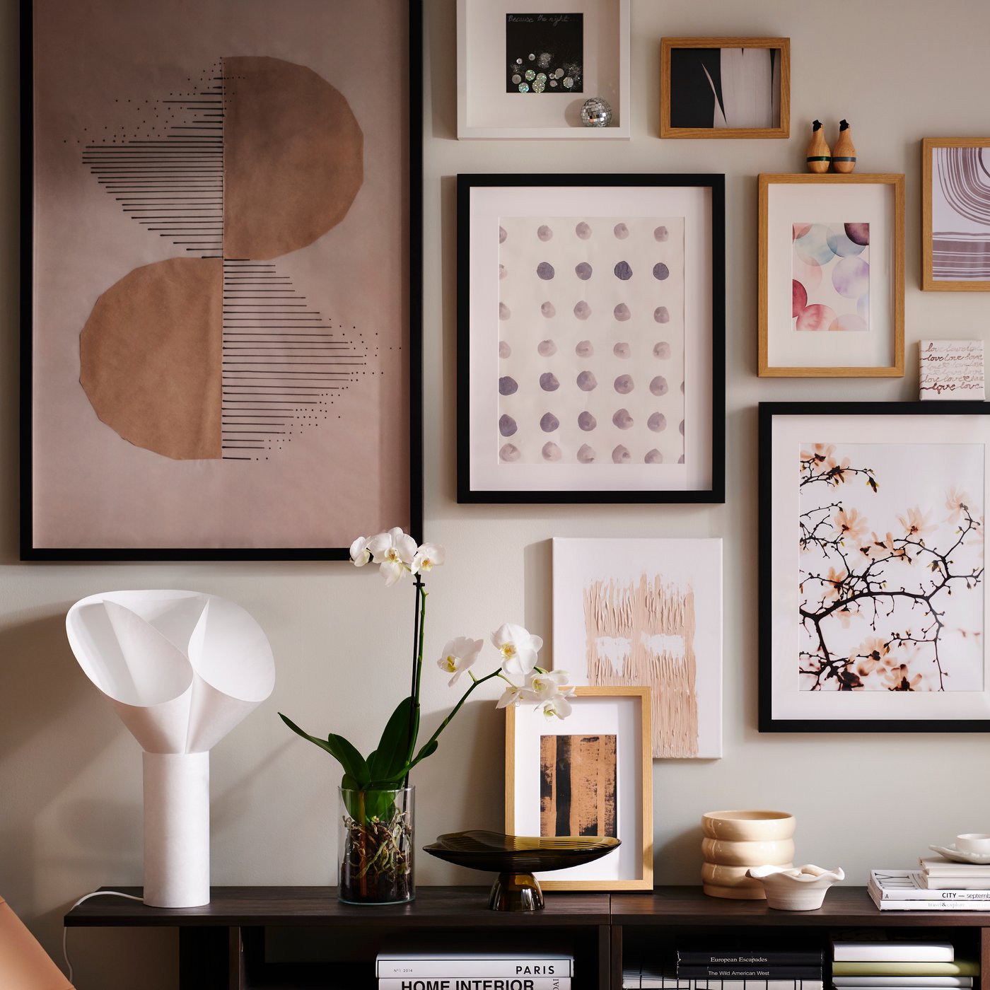 Una parete di un soggiorno luminoso con un gruppo di immagini in varie cornici RÖDALM sopra tavolini HOLMERUD con orchidee e oggetti decorativi.