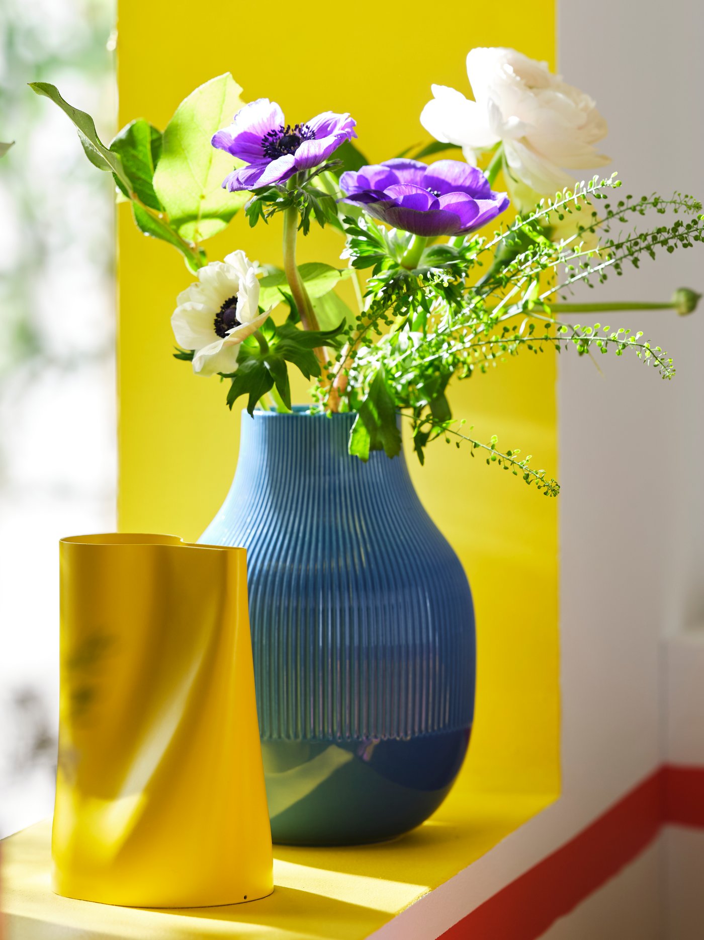 Žuta CHILIFRUKT kantica za zalivanje nalazi se pored plave GRADVIS vaze s cvećem na simsu.