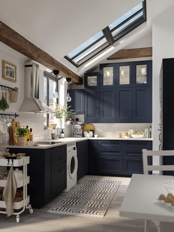 L shaped AXSTAD blue shaker kitchen with wall cabinet with glass doors, white worktop and metal handles.