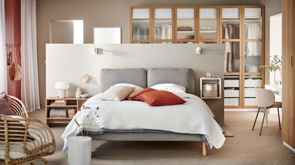 A SAGESUND upholstered bed in Diseröd brown/Luröy in a large airy bedroom covered in white bed linen with red cushions on it.