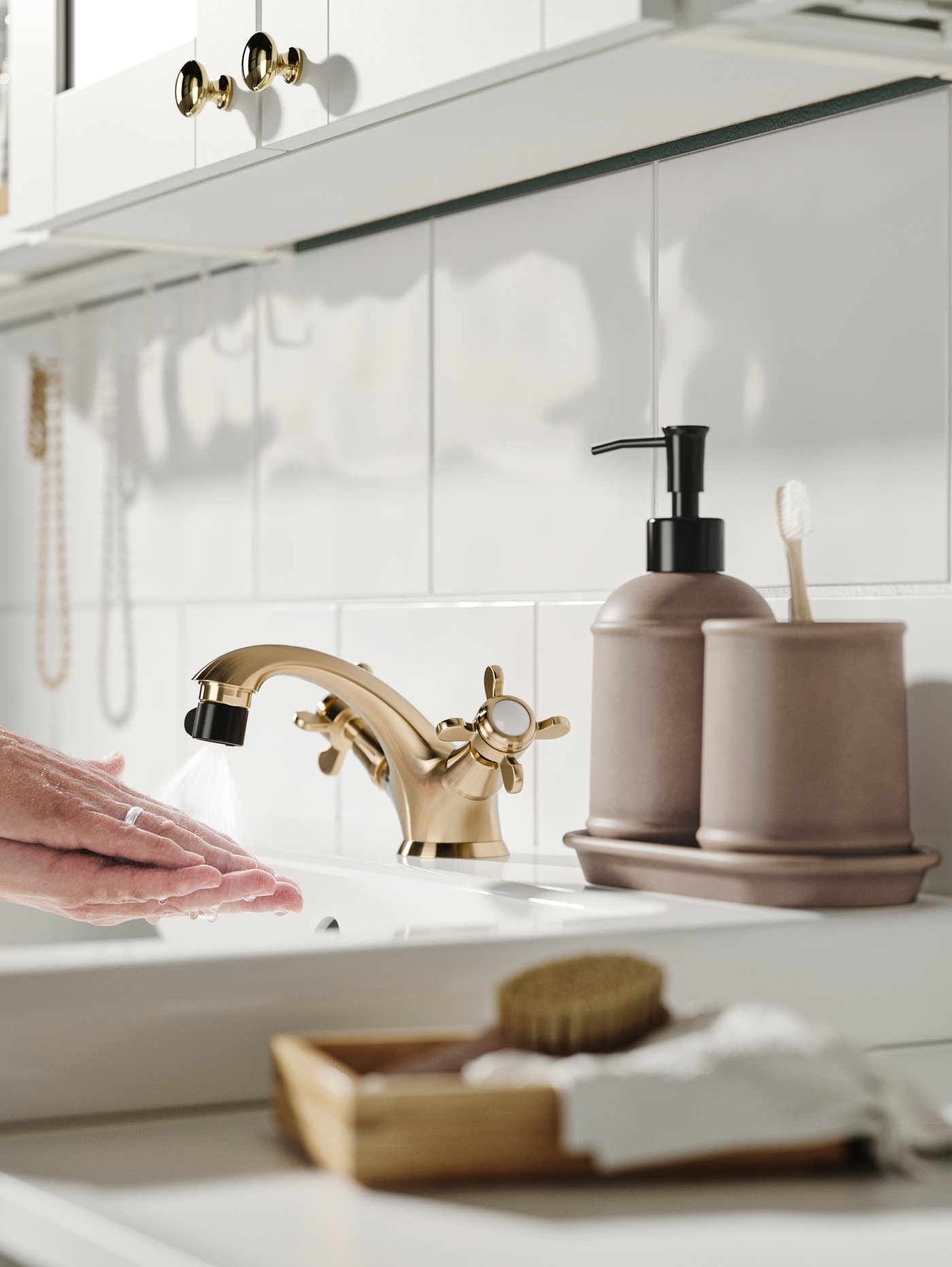 Händer under rinnande vatten i ett badrum med guldfärgad kran, två tandborsthållare och en tvålpump.