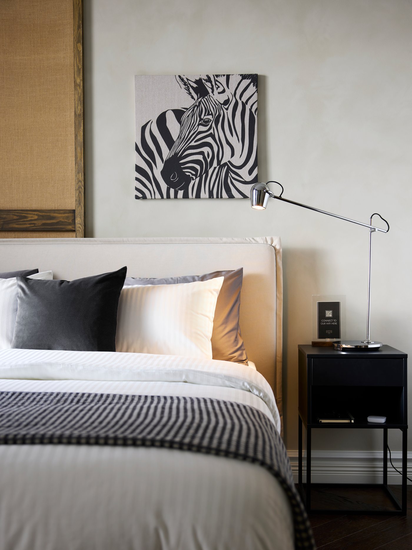 A beige TÄRNKULLEN upholstered bed frame accents a freshly made hotel bed on a wall below a zebra artwork, by a nightstand.