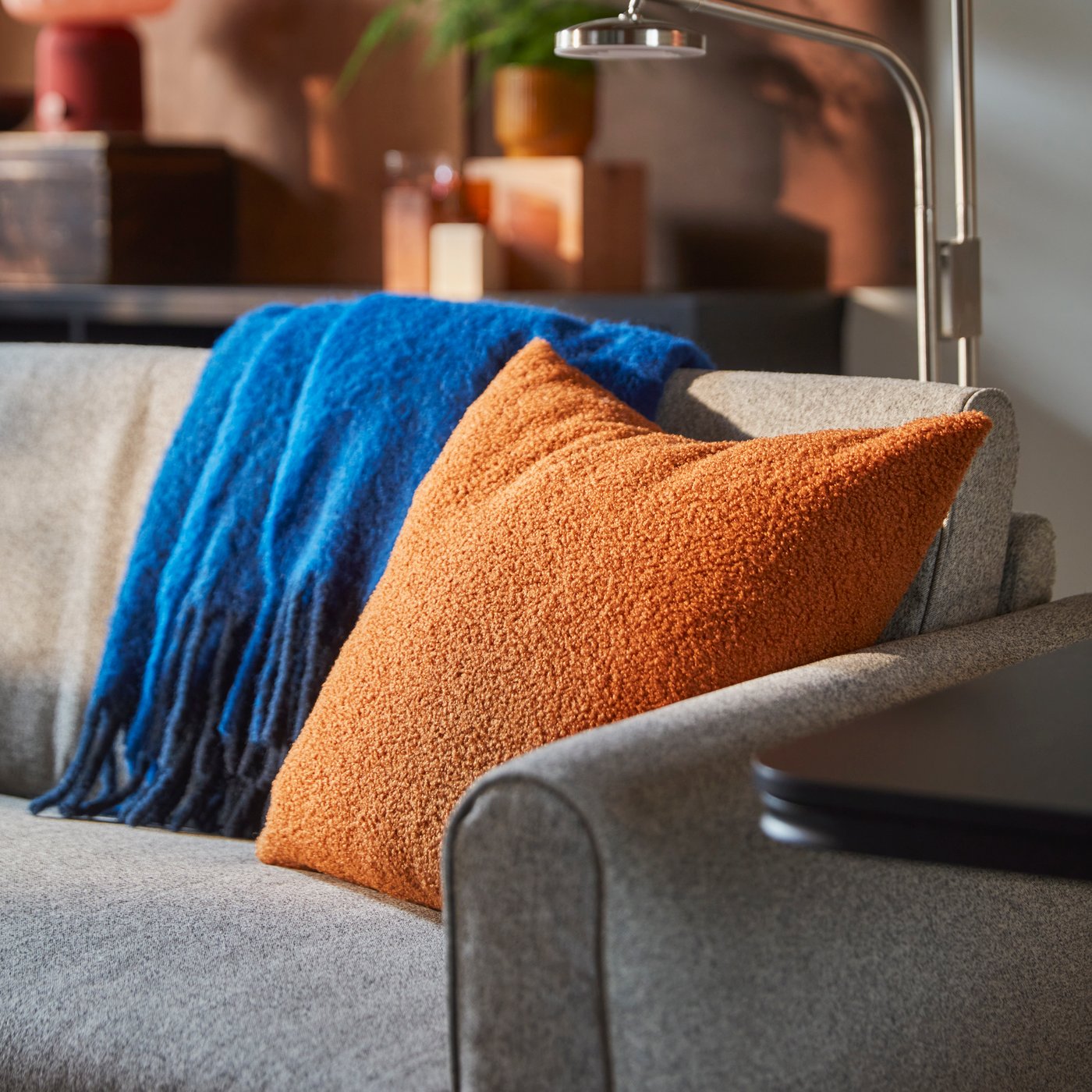 KRYDDBUSKE brown/red cushion cover with a blue throw on a grey sofa.
