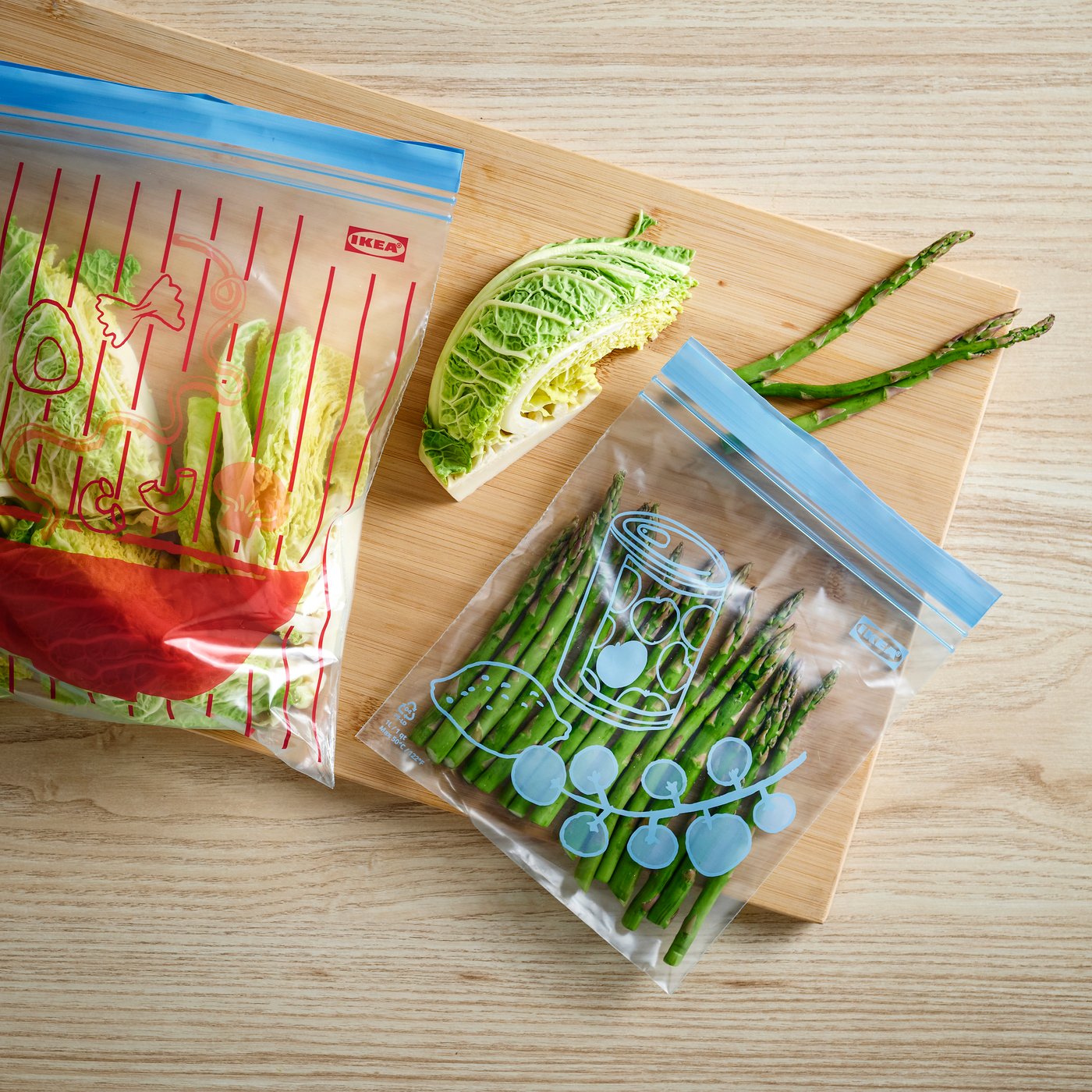 Два прозрачных многоразовых пакета с застёжкой на деревянной доске, в каждом — свежие продукты: салат и спаржа.