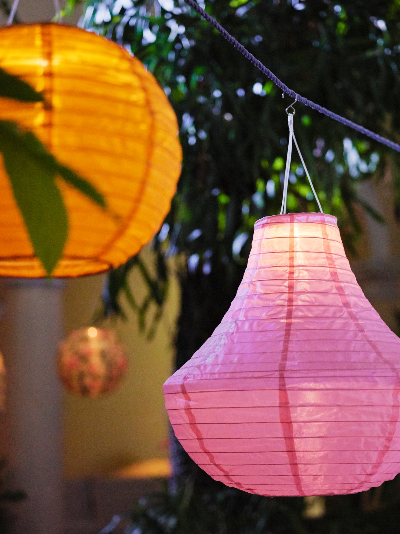 Two lit solar-powered SOLVINDEN pendant lamps in pink and orange hang on a rope outside at night-time.
