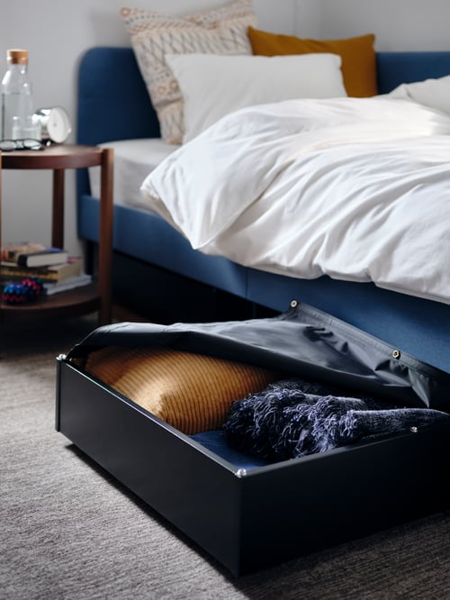 A partially open storage box with a cushion and a throw is next to an upholstered bed frame with a duvet set and pillows.