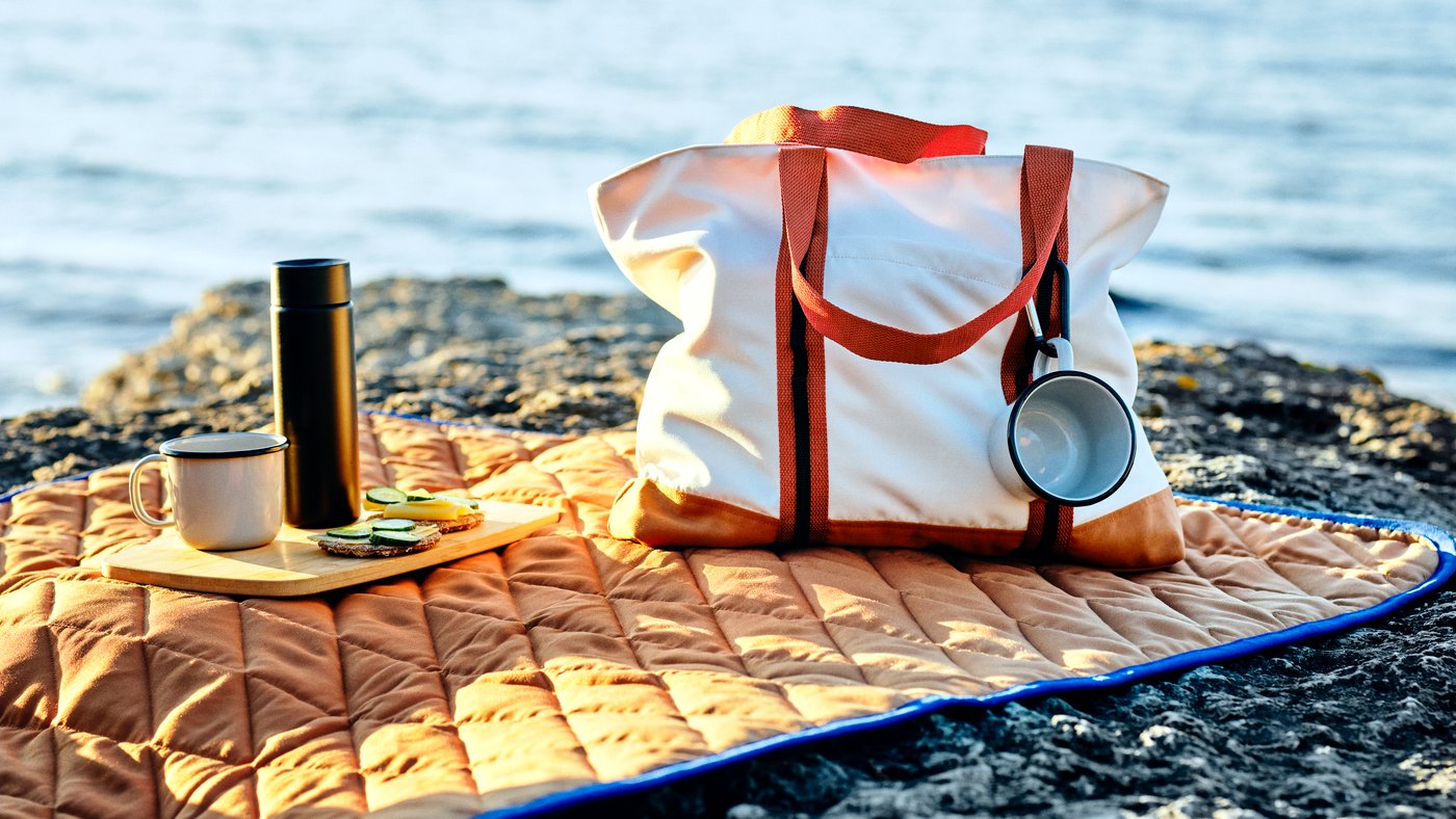 A SOLUPPGÅNG bag, two mugs and a chopping board are on a SOLUPPGÅNG picnic blanket on a rocky cliff by the sea.