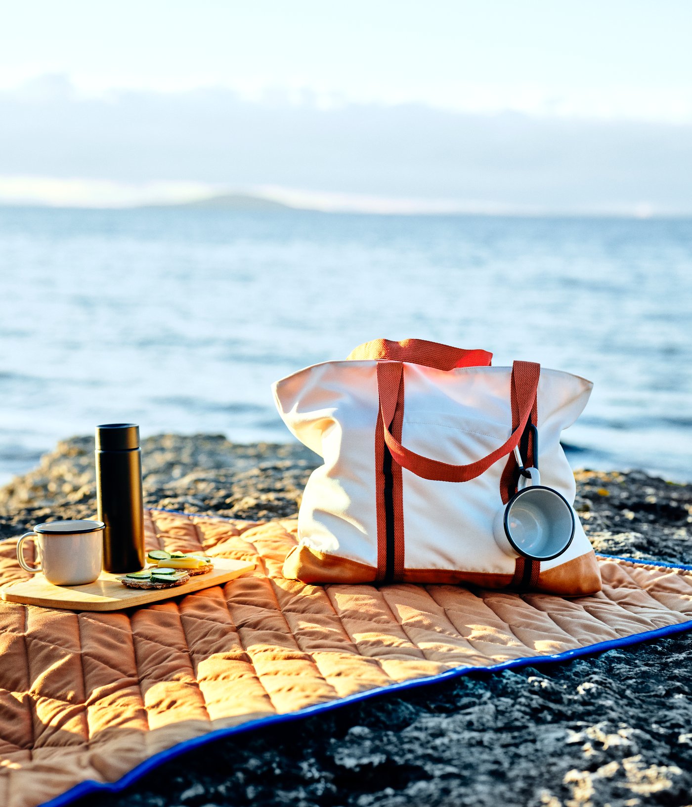 A SOLUPPGÅNG bag, two mugs and a chopping board are on a SOLUPPGÅNG picnic blanket on a rocky cliff by the sea.