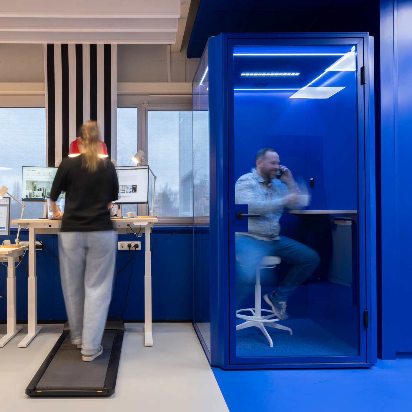 Une cabine téléphonique bleu vif, à côté d’un poste de travail IKEA debout, crée un coin pratique pour des appels concentrés, tandis qu’une personne voisine travaille activement sur un bureau avec tapis de marche.
