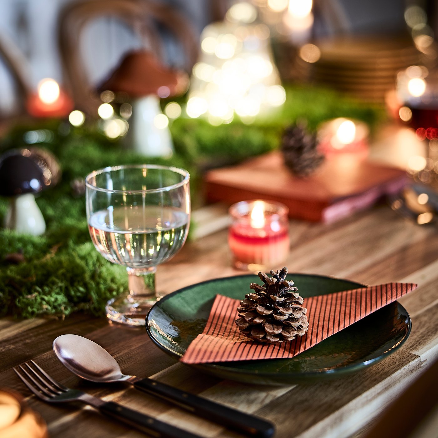 An autumn-themed table setting with a pinecone resting on a folded HÖSTAGILLE paper napkin placed on a green glass plate.