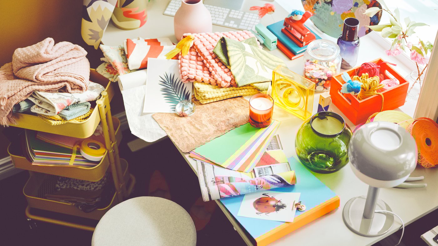 A desk is covered with colourful cushion covers, paper and a green vase. A yellow RÅSKOG trolley is to the side.