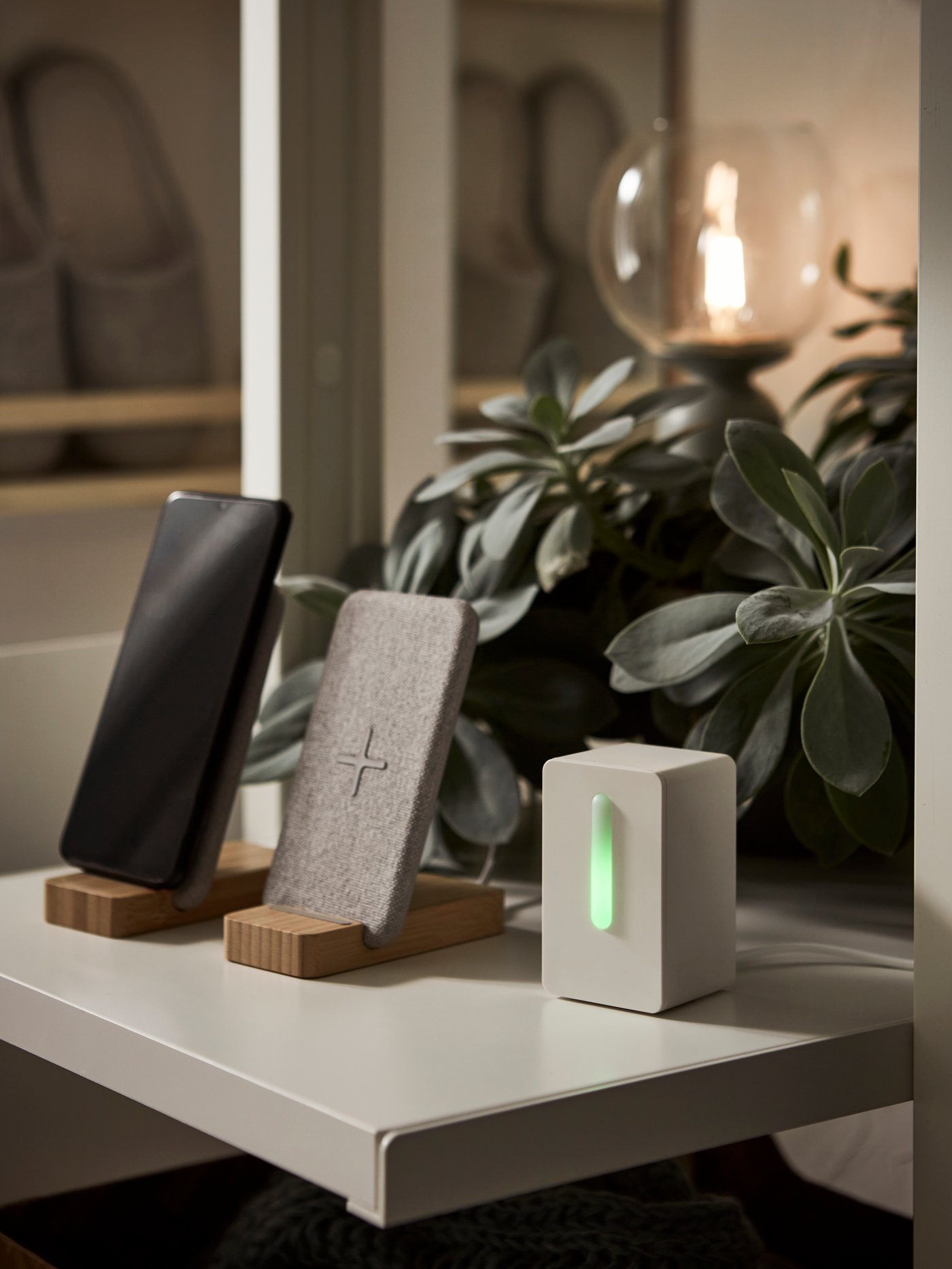 Na polici so VINDRIKTNING senzor kakovosti zraka in dve NORDMÄRKE stojali za brezžično polnjenje, v ozadju pa bujna zelena rastlina.