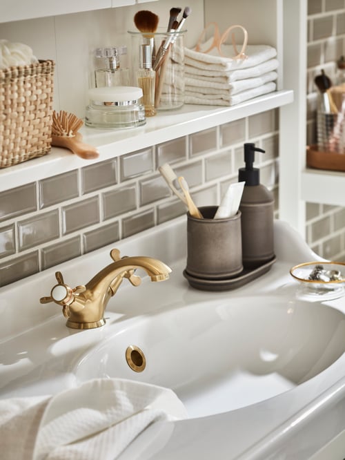 Close-up on bathroom rectangular ceramic wash basin with deep water trap, gold coloured faucet, and accessories on the sides.
