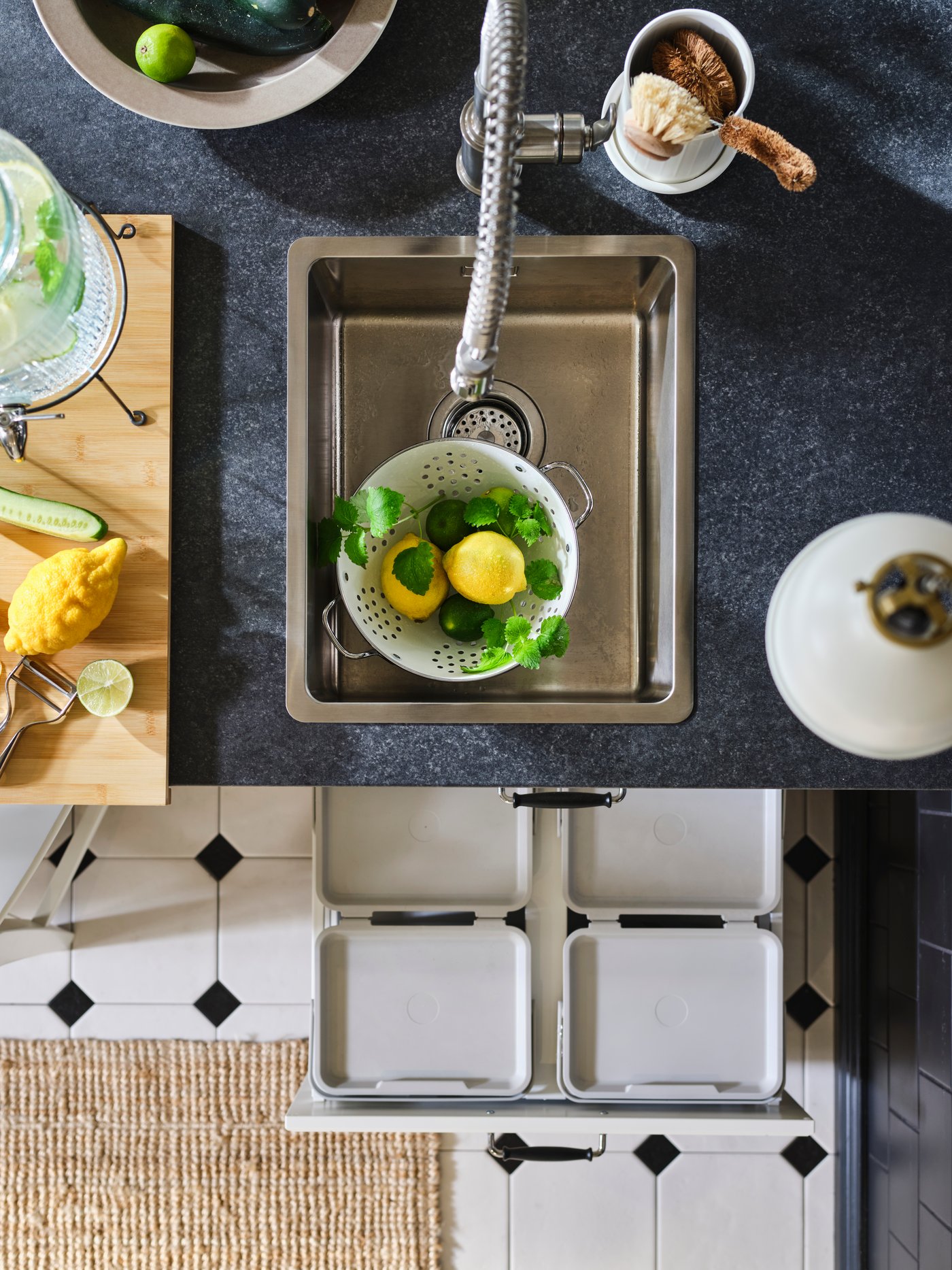 A black kitchen worktop with VRESJÖN insert sink in stainless steel, holding colander filled with lemons and herbs.