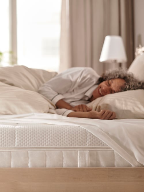 A person lays on a foam mattress with a duvet covering them, and a fitted sheet partially covering the mattress.