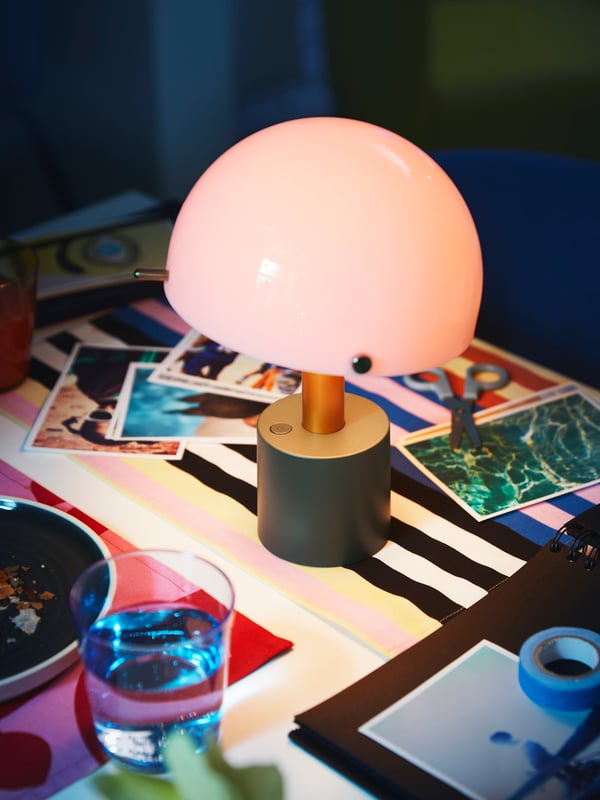 A light-pink-and-dark-grey-green NÖDMAST LED portable lamp lights up a tabletop covered in colourful textiles.