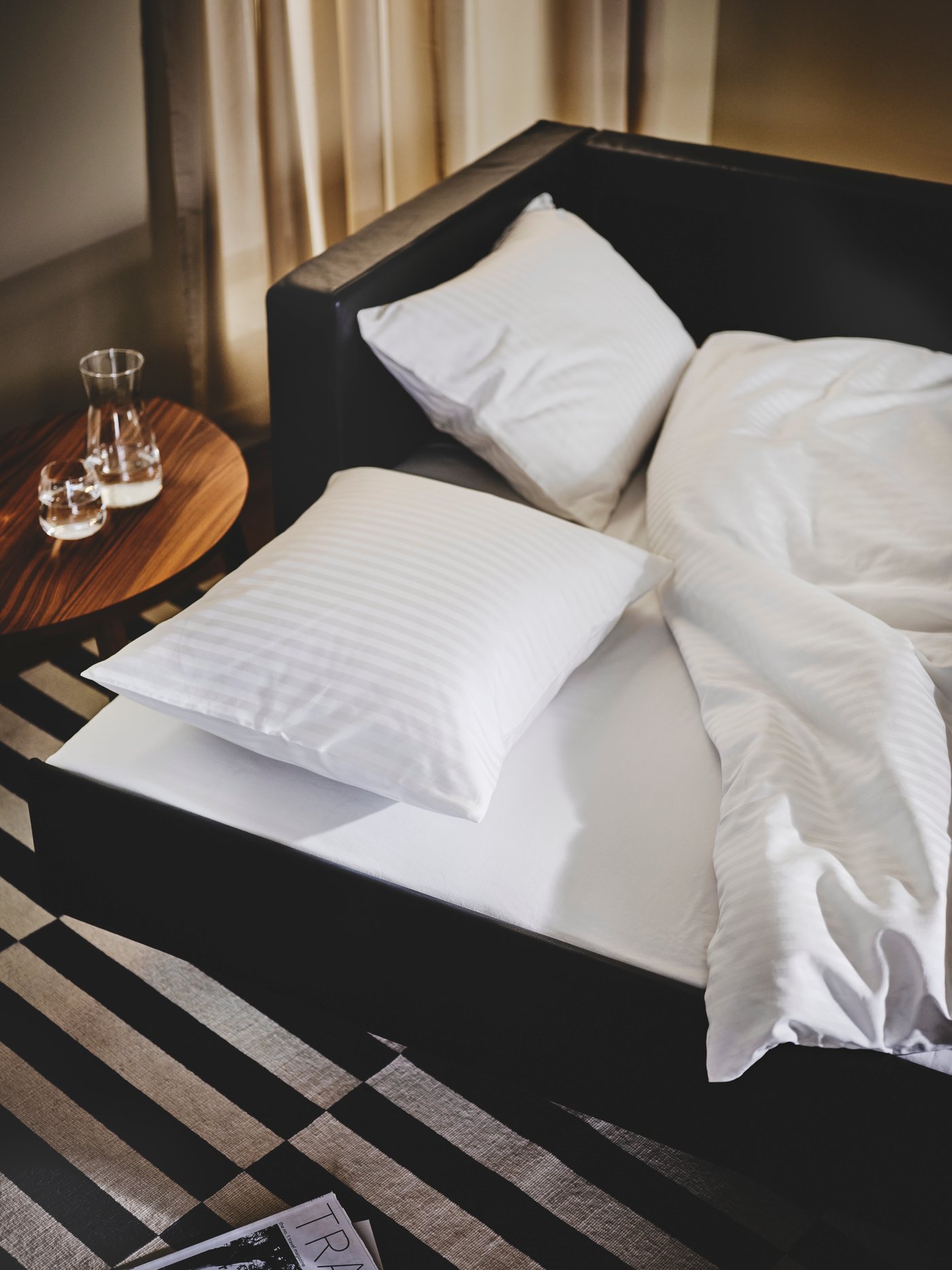 Primer plano de un sofá cama LANDSKORNA convertido en cama con un juego de edredón NATTJASMIN. La cama se encuentra sobre una alfombra de rayas.