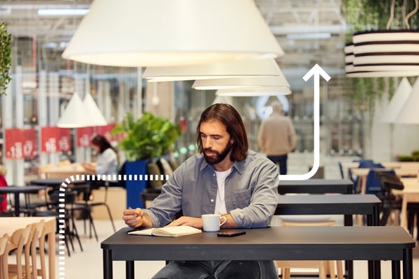 A man sits in an IKEA Swedish restaurant drinking coffee while he writes in a notebook.