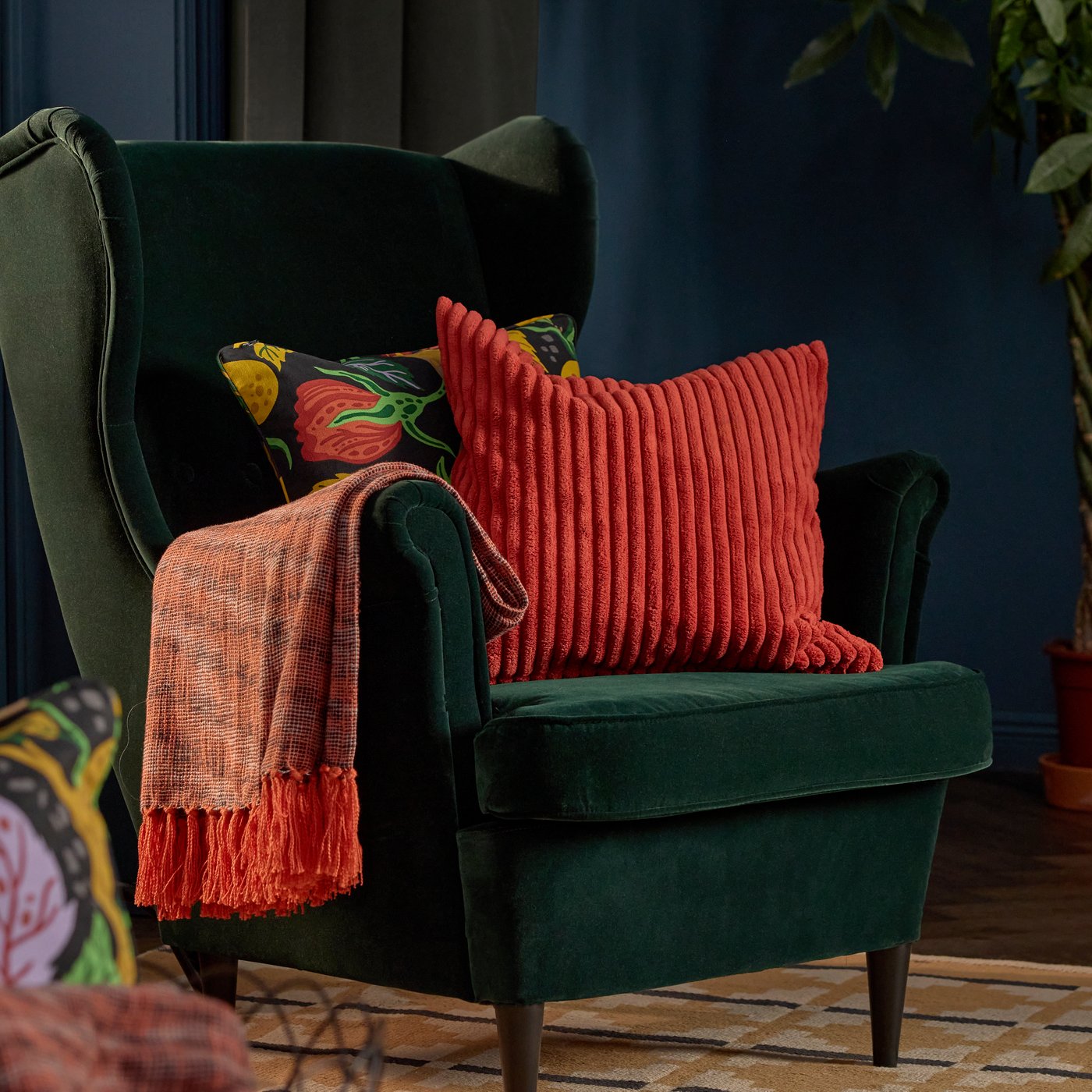 غطاء HÖSTAGILLE مع أطراف متدلية بلون برتقالي مصنوع من قطن 100%، معلق على ذراع كرسي بذراعين أخضر غامق عليه غطاءين وسادة بنمط يتماشى مع فصل الخريف.