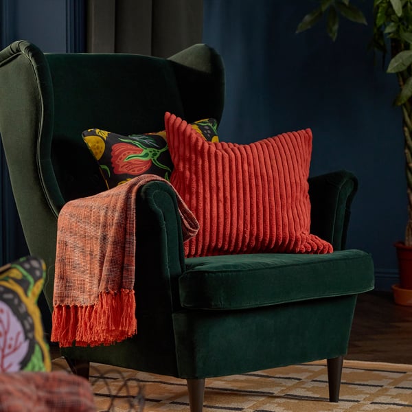 Un plaid HÖSTAGILLE arancione in 100% cotone, con le frange, è appoggiato sul bracciolo di una poltrona verde scuro con due fodere per cuscino in colori e motivi autunnali.