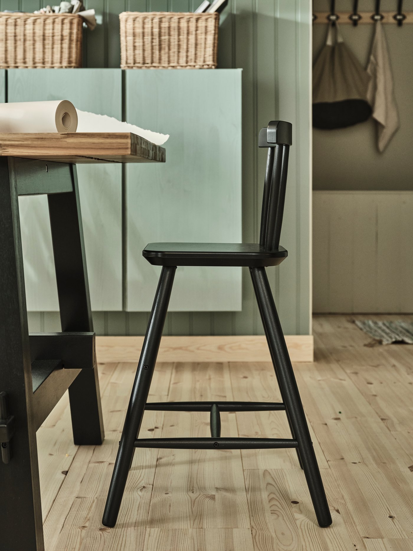 A black AGAM junior chair next to a SKOGSTA table with acacia wooden top and black legs and a roll of art paper on top.