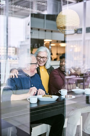 Trei persoane stau la o masă în restaurantul IKEA, văzute prin geam, se bucură de mâncare și cafea într-un spațiu luminos, cu atmosferă caldă și relaxată.