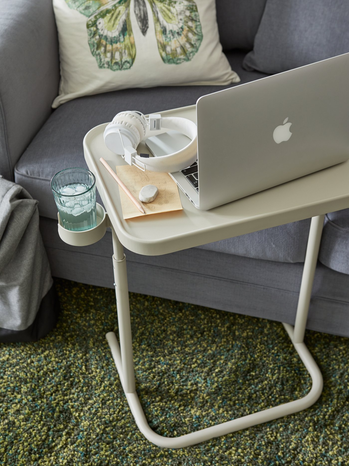 Ein beigefarbener Laptophalter vor einem grauen Sofa mit einem Laptop, weißen Kopfhörern, Bleistift, Radiergummi und einem Glas Wasser.