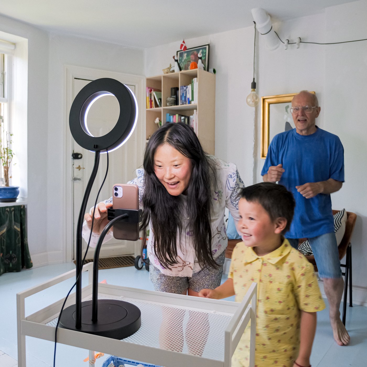 Una mamma e il suo bambino si preparano a registrare un video sistemando una luce ad anello LÅNESPELARE con supporto per cellulare. Dietro di loro il nonno balla.