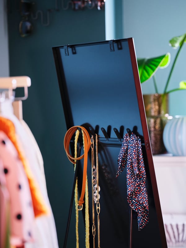 The back of a black KARMSUND standing mirror showing hooks holding belts and a scarf, with clothes on hangers beside it.