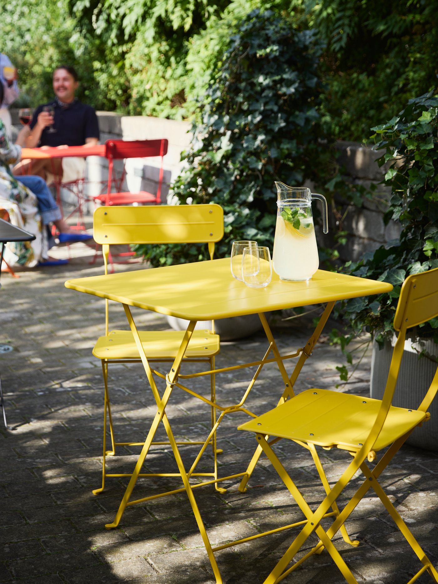 Bright yellow SUNDSÖ table in a sunny courtyard, with people enjoying their time in the background.