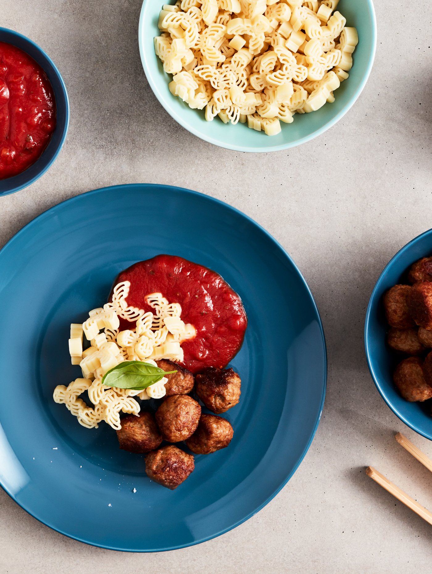 A dark turquoise plate and bowls and one green bowl, holding HUVUDROLL meatballs, BÄSTISAR pasta and tomato sauce.