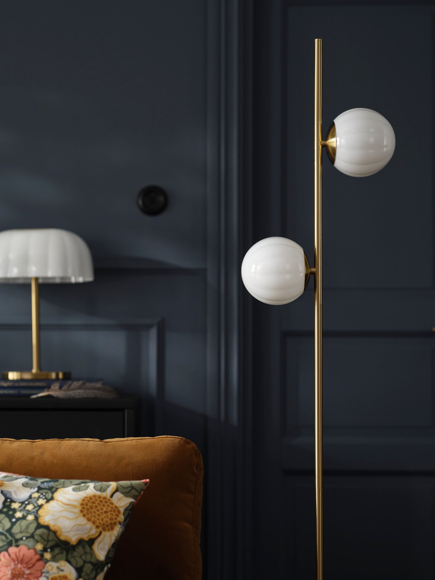 An AKTERSPRING floor lamp in opal white glass and brass-plated steel stand in a living room with a dark blue wall and a sofa.