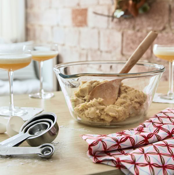An IKEA VARDAGEN glass bowl filled with mixing dough, a wooden mixing spoon, stainless steel measuring cups, a white and red tea towel on a light oak surface and cocktail glasses in the background.