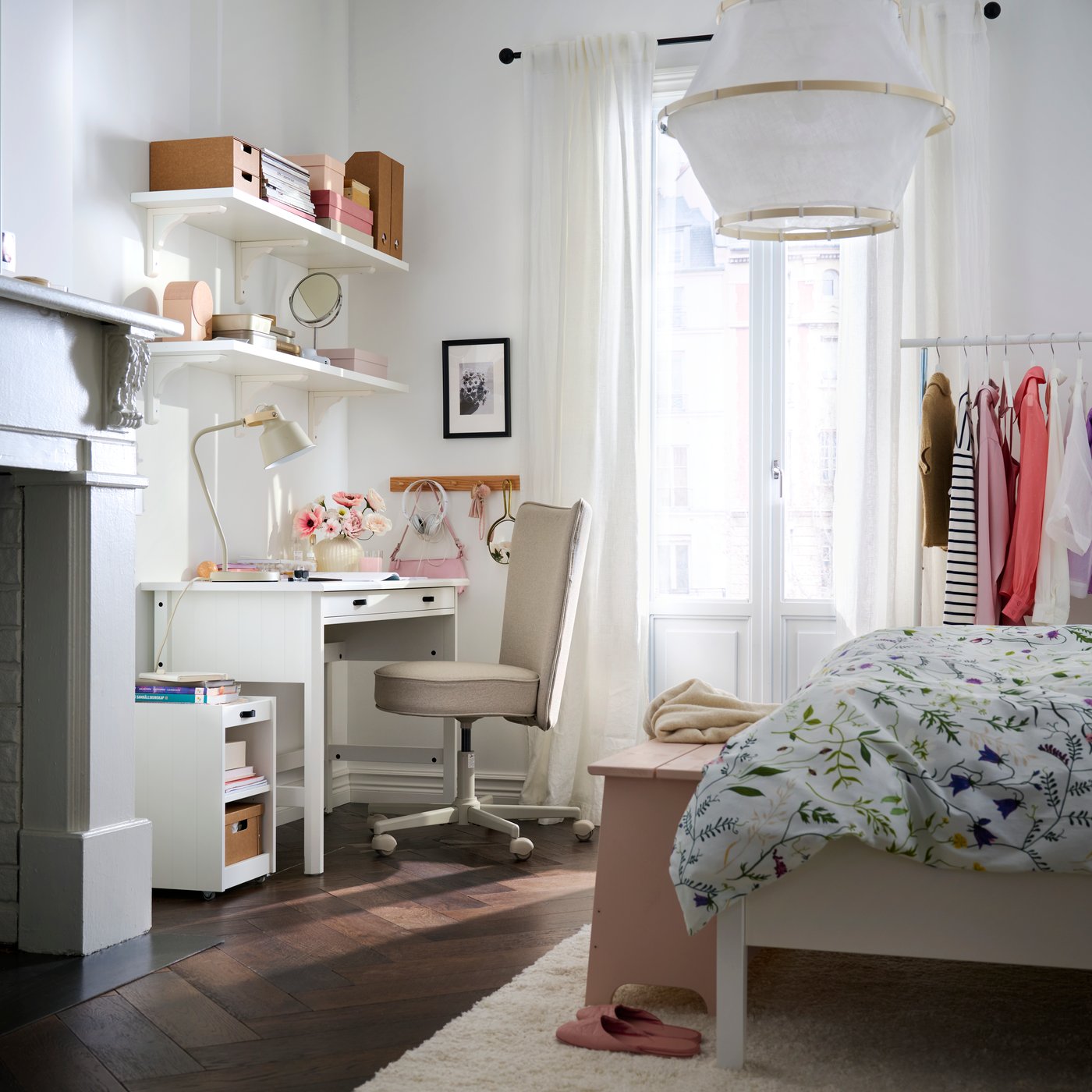 A bedroom with a white BRYTNING desk made from white material sits beside a bed with floral bedding and clothing racks.