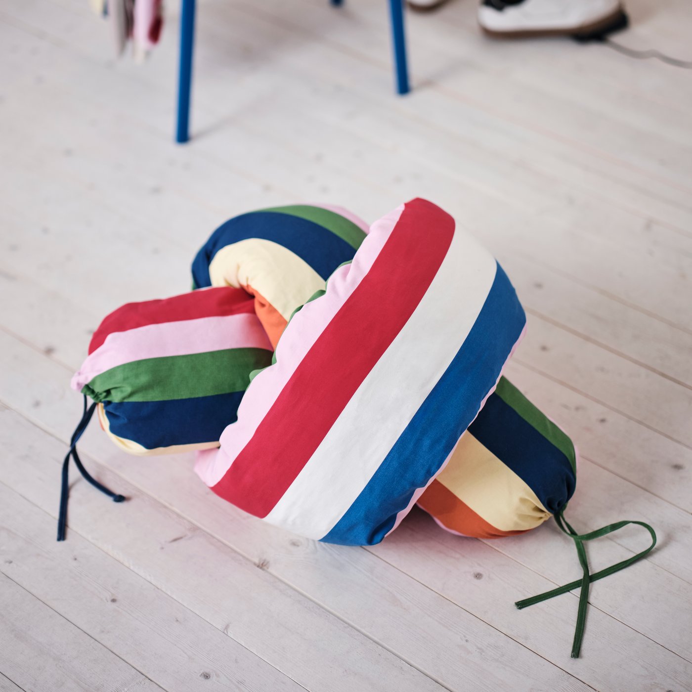 A tie-knot pillow lies on a bright wooden floor. It’s made with striped, candy-coloured JÄTTEVÄDD pre-cut fabric.