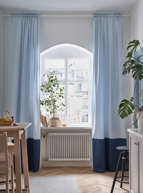 Køkkenvindue med en stor plante i vindueskarmen og lyseblå BENGTA mørklægningsgardiner på begge sider.