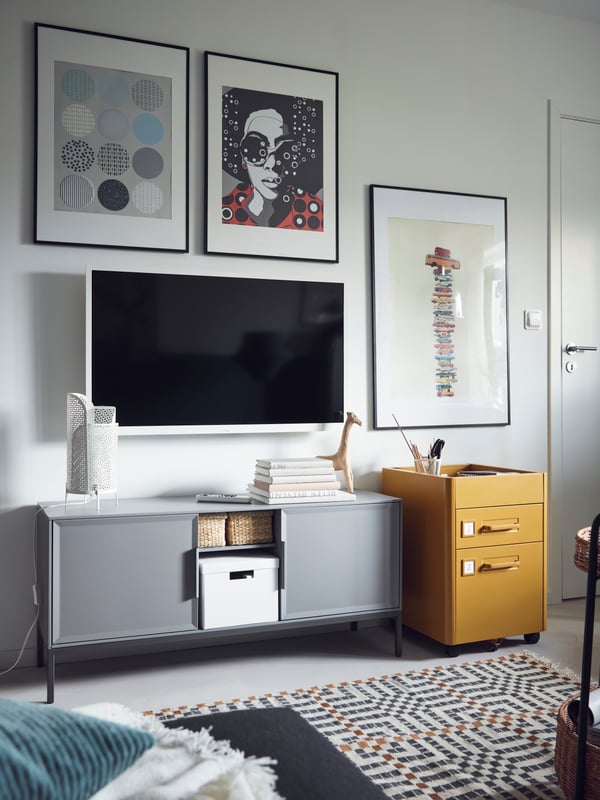 Een tv en foto’s hangen aan de wand boven een grijze TULLSTORP metalen kast met open opbergruimte, met manden en een doos erin.