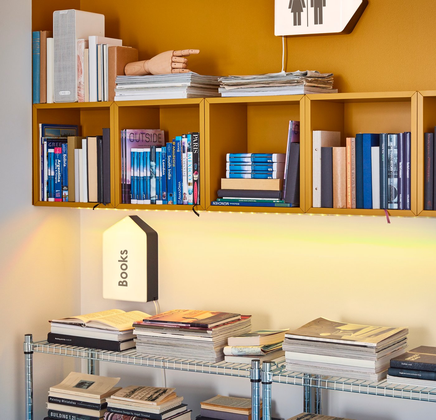 Ein modernes Bücherregal mit offenen, senfgelben Wandwürfeln, gefüllt mit Büchern und Zeitschriften, darüber ein leuchtendes Wandbild mit WC-Symbolen; darunter ein Wandlicht in Form eines Hauses mit der Aufschrift „Books“ sowie ein Metallregal voller aufgeschlagener und gestapelter Bücher, vor einer warm beleuchteten Wand.