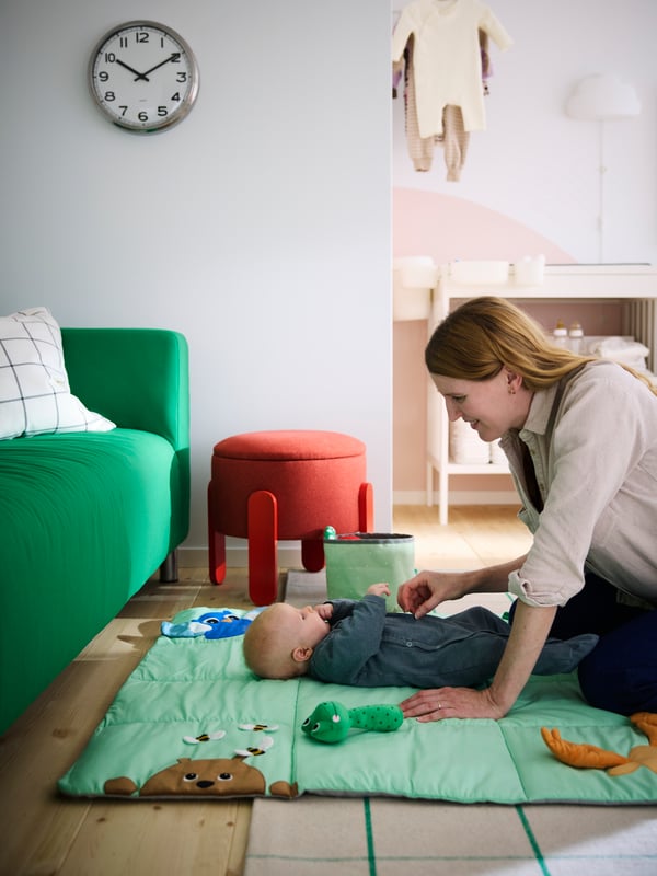 En mamma kittlar sin bebis som ligger på en ljusgrön SANDBI lekmatta, bredvid en grön soffa och med en röd sittpuff i bakgrunden.