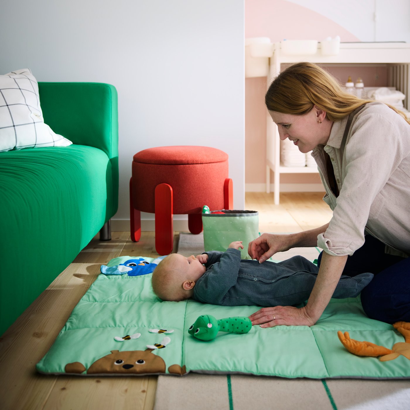 A mom tickling her baby laying on a bright green SANDBI play mat, next to a green sofa and with a red pouffe in the back.
