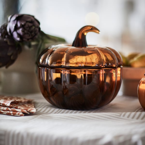 A close-up of the yellow-brown HÖSTAGILLE decorative bowl with a lid, featuring a pumpkin-shaped design on a patterned table.