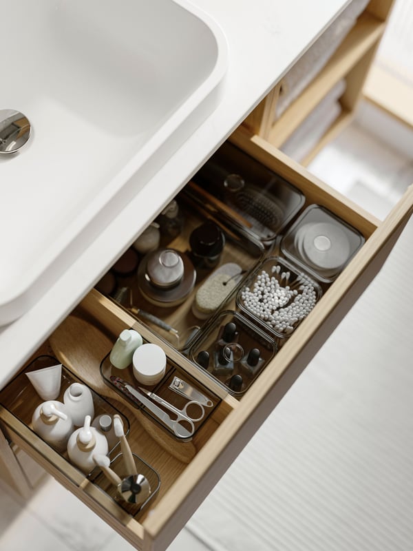 A bathroom vanity has a drawer that’s open and there are care products organised in grey VISSLAÅN boxes with lids inside.
