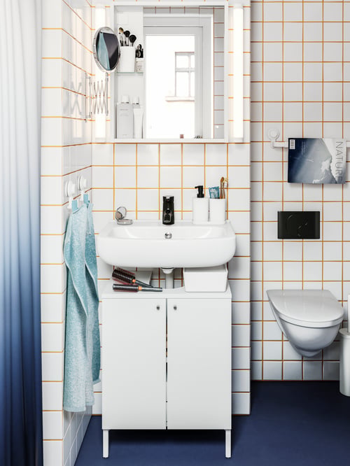 Ein weiß gefliestes Badezimmer mit einem NYSJÖN Waschbeckenunterschrank, einem FRÄCK Spiegel, TISKEN Haken mit Saugnäpfen und zwei Handtüchern.
