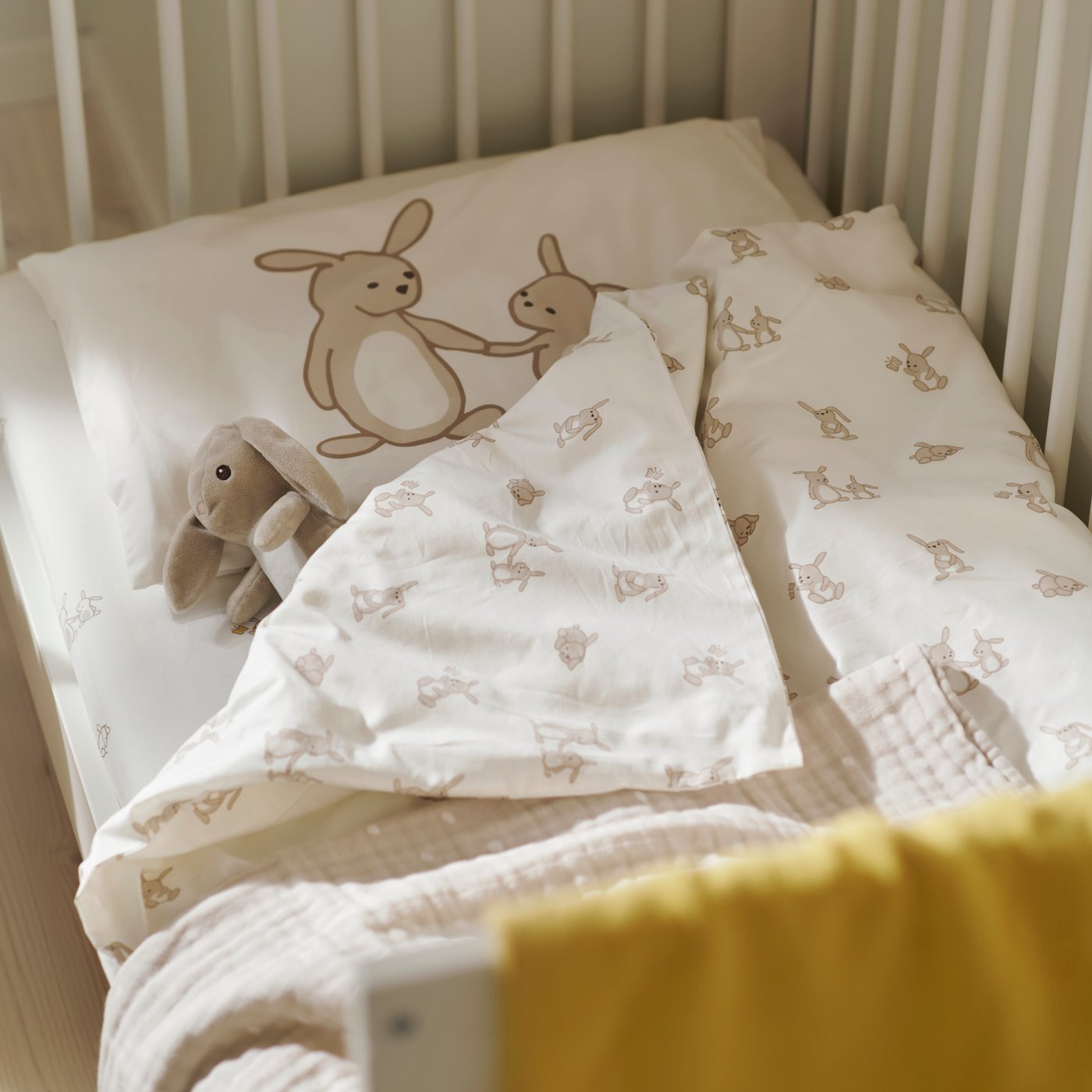 A LEN blanket in white/beige with a texture on a cot, alongside rabbit-print bedding and a yellow blanket in a nursery.