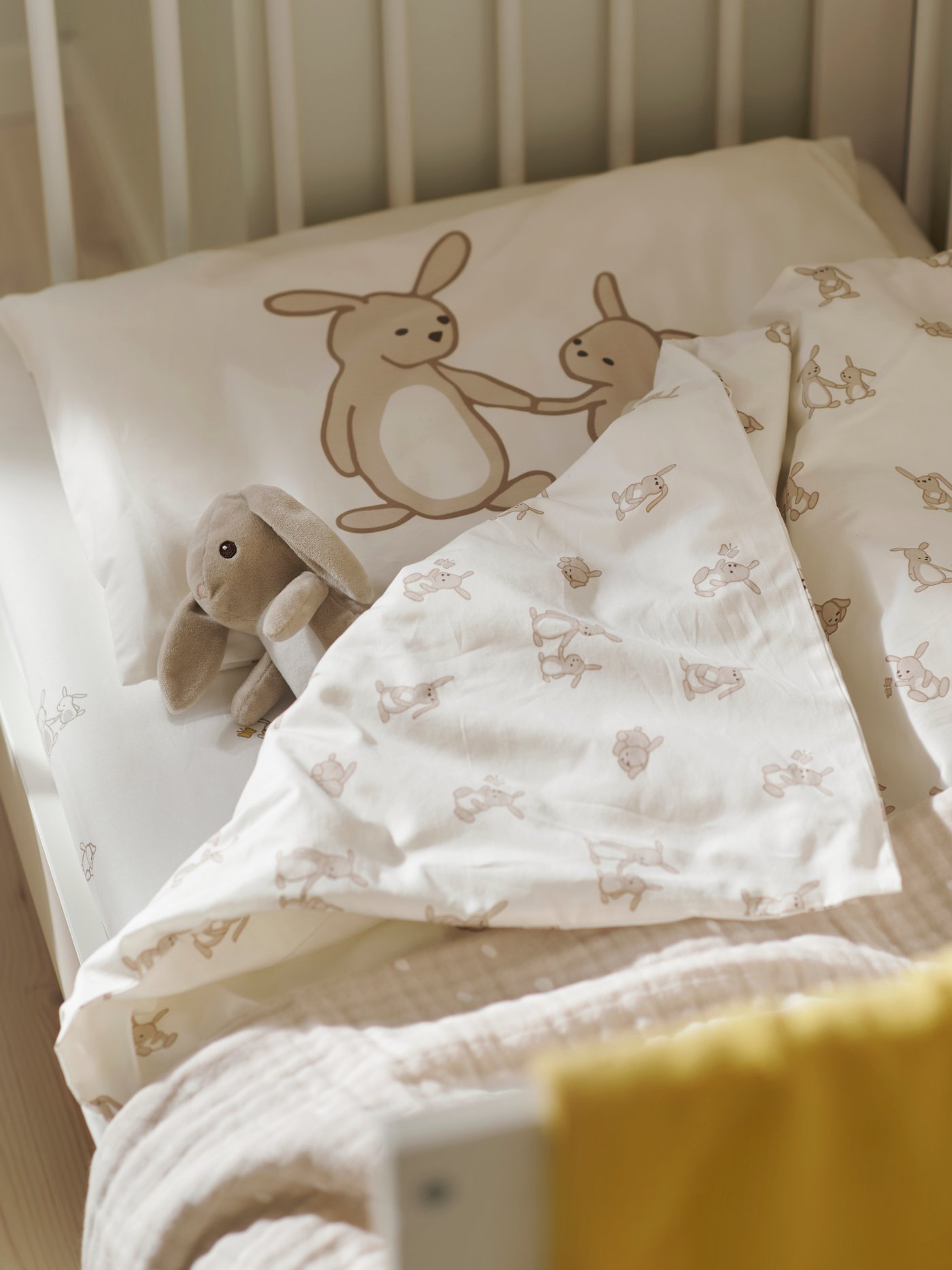 A LEN blanket in white/beige with a texture on a cot, alongside rabbit-print bedding and a yellow blanket in a nursery.