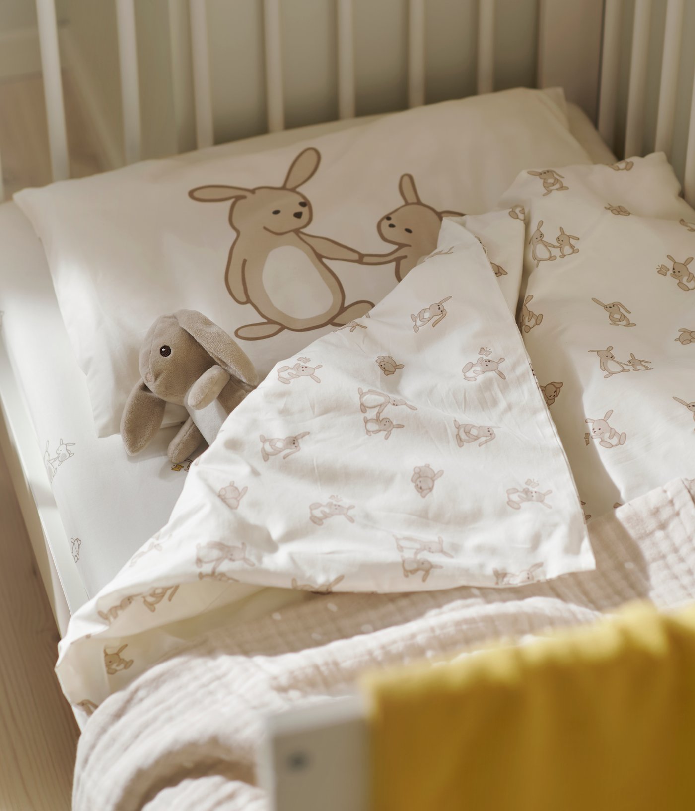 A LEN blanket in white/beige with a texture on a cot, alongside rabbit-print bedding and a yellow blanket in a nursery.