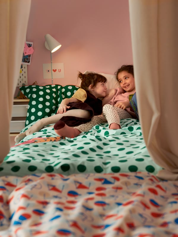 Two young girls relaxing in a bed made with a colorful green dotted quilt cover placed in a colorful children’s room.