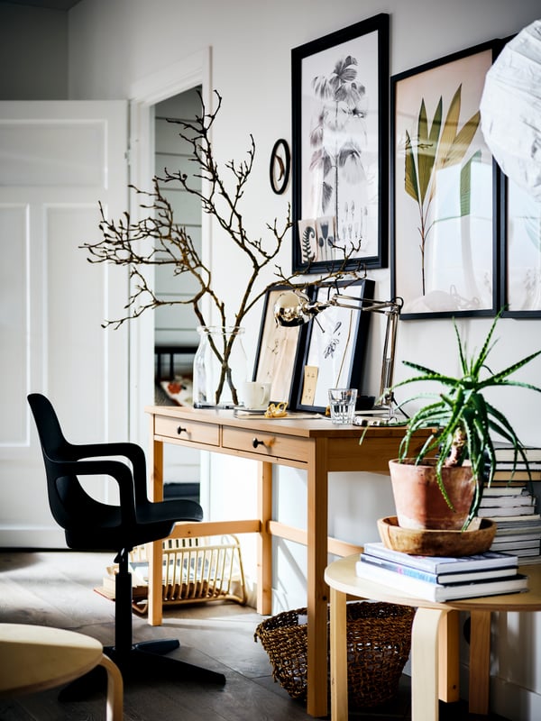 A HEMNES desk with a vase of branches on top, against a white wall covered with pictures. A swivel chair is in front.